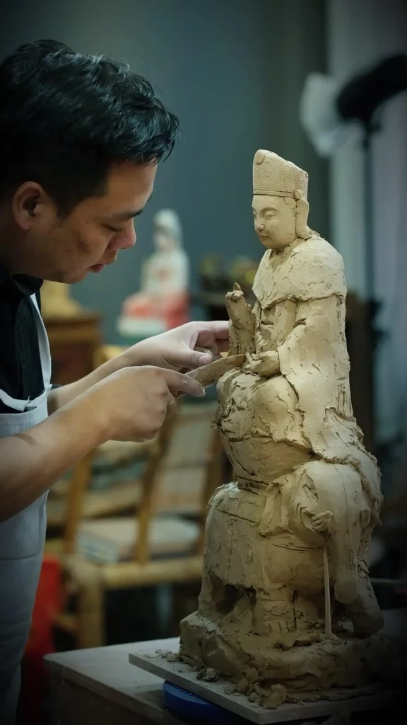 A sculptor shaping the hands and robes of a clay figure.