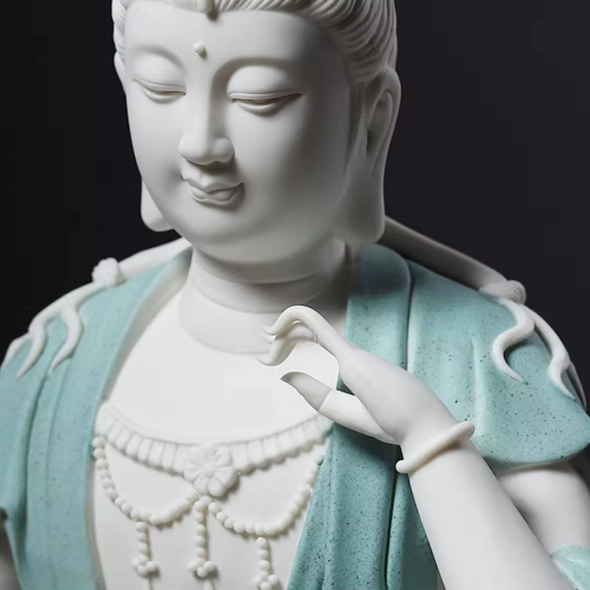 Close-up of the contemplative Kuan Yin statue’s lowered eyes, curved raised fingers, cream-white necklace and finely grained teal robe