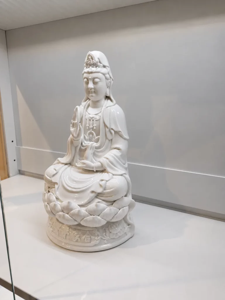 Customer photograph of a white porcelain Guanyin on a lotus seat in a glass cabinet, front view.