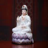 Opposite angled view of a seated Guanyin statue with white robes, gold-colored borders and a purple lotus base