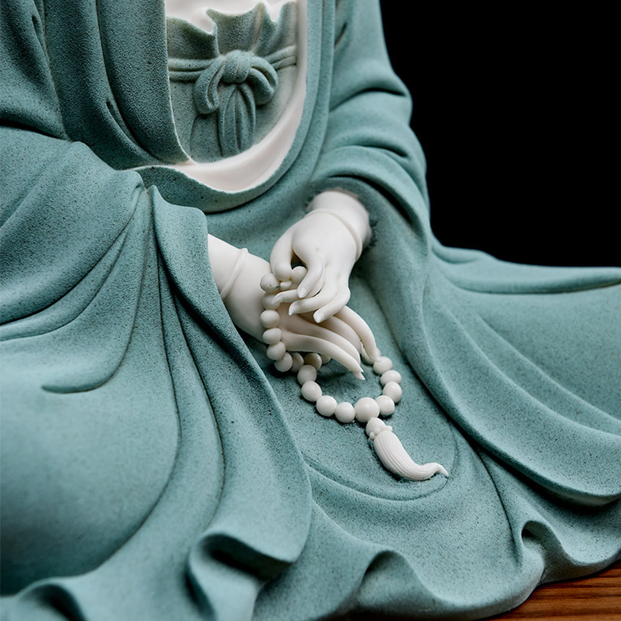 Close-up of the teal Jingsi Guanyin statue’s hands holding sculpted prayer beads and a tassel over textured robes